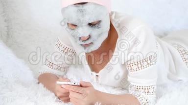一个躺在沙发上的年轻女人用她的智能<strong>手机</strong>上网。 女人用泡泡面<strong>膜</strong>敷脸皮